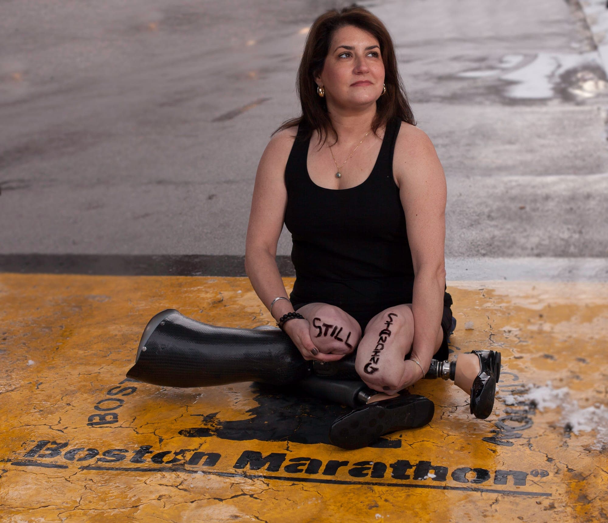 Celeste Corcoran seated at the Boston Marathon finish line with 'Still Standing' written on her legs, prosthetics beside her