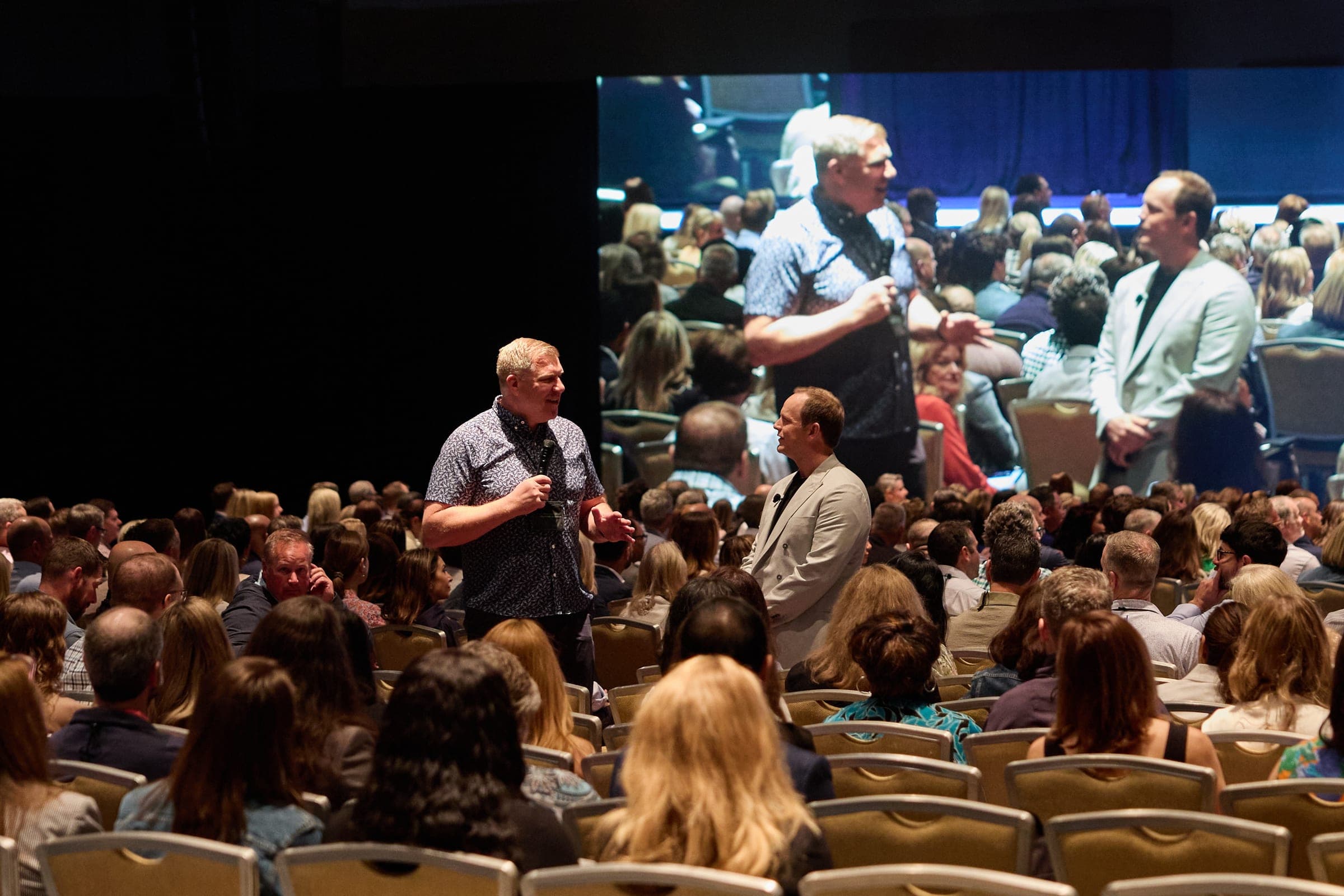 An audience member stands with a microphone in the middle of a seated ballroom, telling his story during a Dear World keynote.