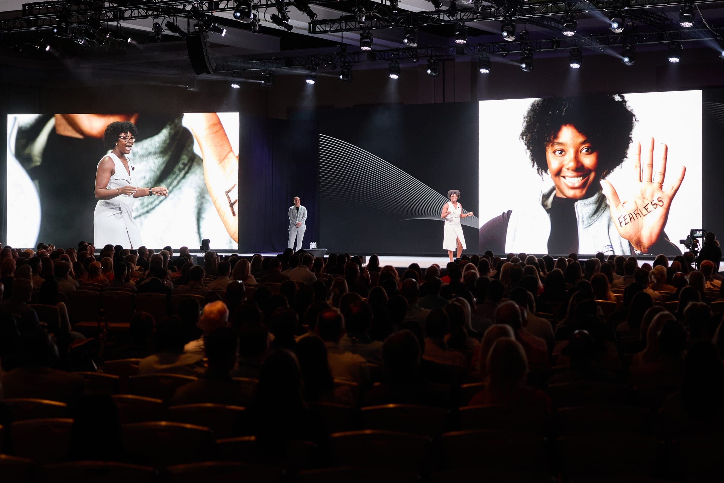 A participant tells her story from the stage while her Dear World portrait fills the screen behind her — the word FEARLESS written across her palm.