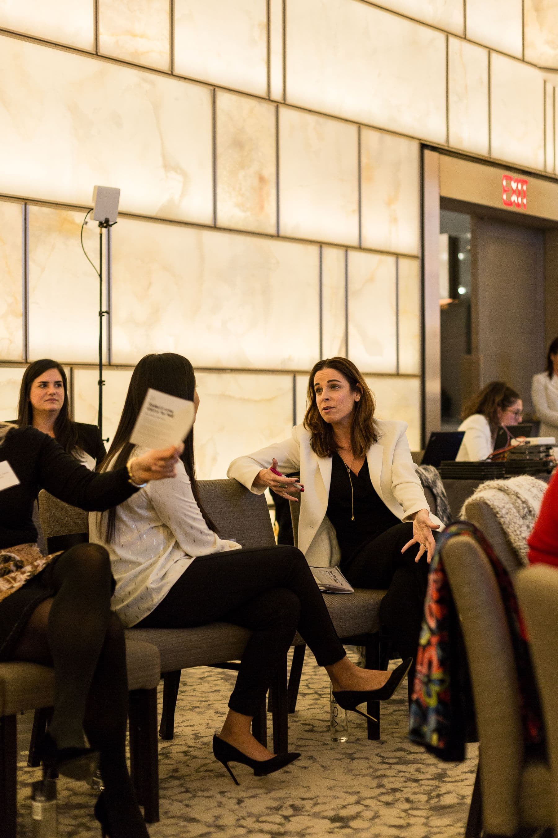 Attendees seated in a small-group conversation at a corporate event, one gesturing while holding a printed card